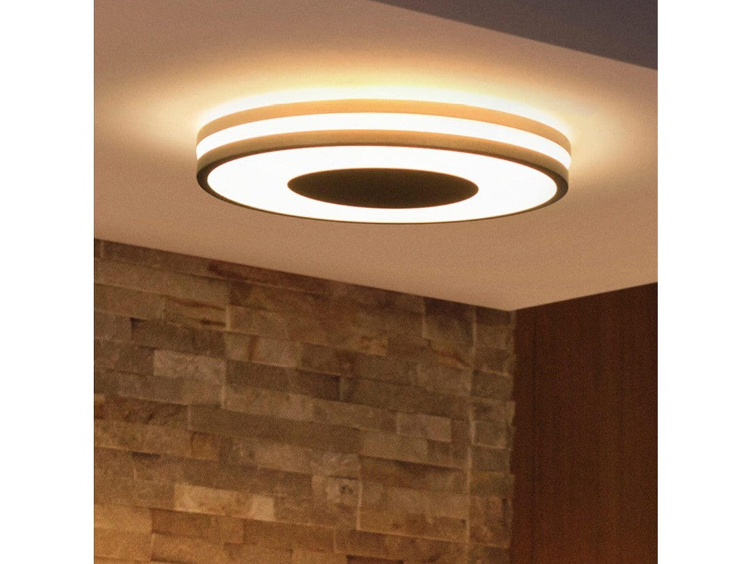 Philips HUE Being loftslampe monteret i loftet med en moderne, cirkulær form og flere lag af lys i varme nuancer over en væg af sten.