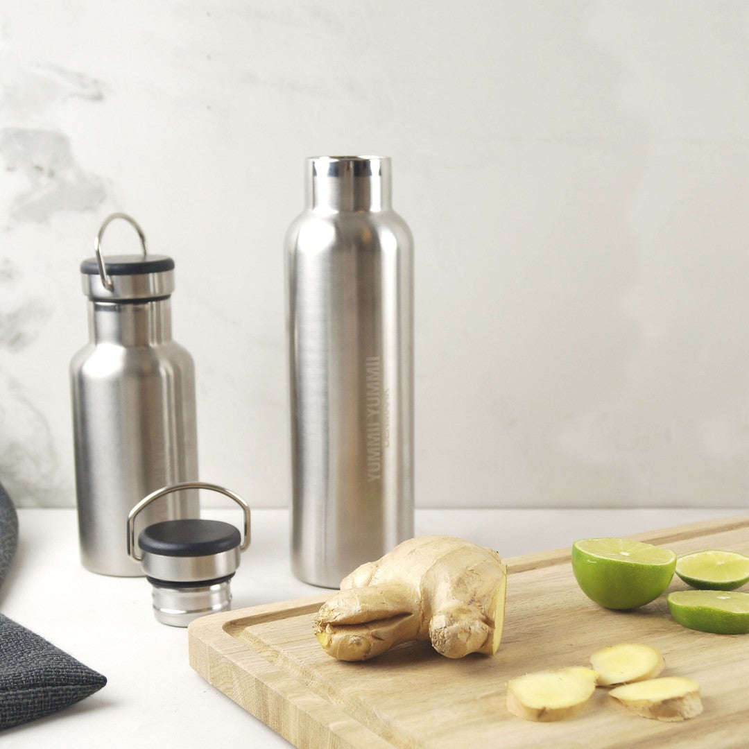 YUMMII YUMMII termoflaske i rustfrit stål på 350 ml i sort farve placeret ved siden af en mindre flasker med låg, ingefærrod og lime skiver på en træskærbræt med grå stof og marmor baggrund