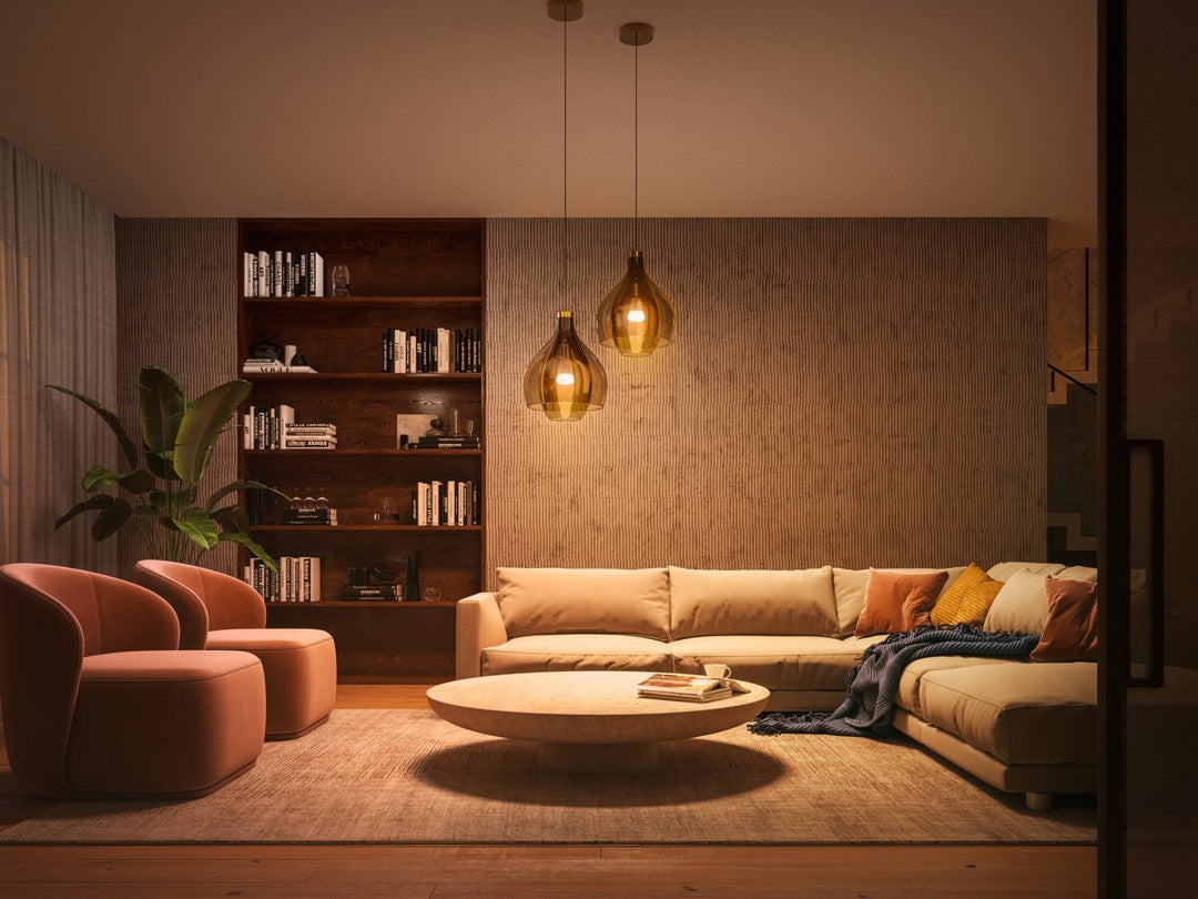 Lysende stue med to rosa lænestole, en beige sofa og et rundt sofabord omgivet af bøger på en reol og en indendørs plante, belysning fra guldpendelstik.