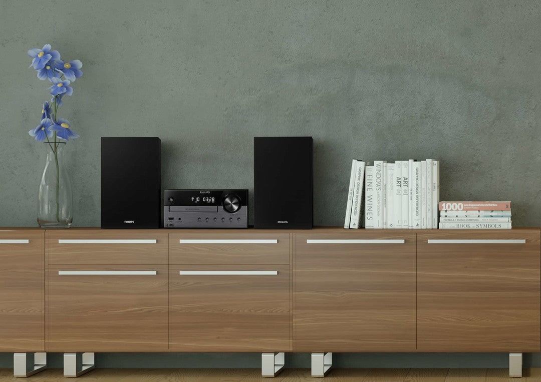 Philips TAM4505/12 mikromusiksystem med to sorte højttalere og en central enhed på et moderne træmøbel med bøger og en vase med blå blomster i baggrunden