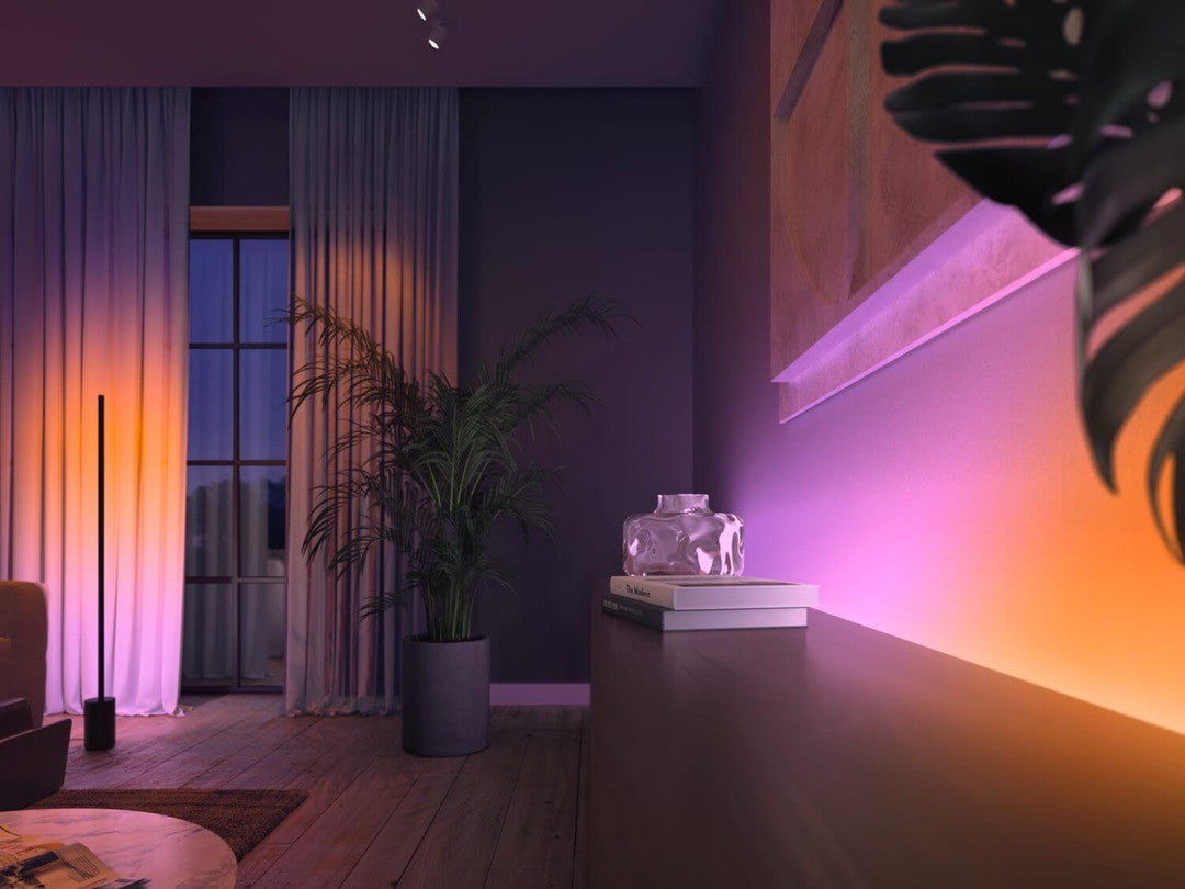 Philips HUE Ambiance gradient lightstrip på 2 meter belyst i et moderne rum med bløde nuancer af orange og lilla der skaber en hyggelig atmosfære planter ved siden af en kommode og vinduer med gardiner i baggrunden
