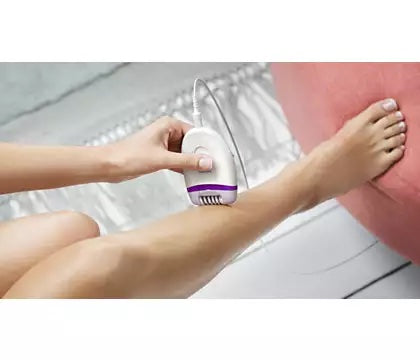 En person der bruger Philips BRE225/00 kompakt epilator på benet mens de sidder på en stol med en rød pude i baggrunden