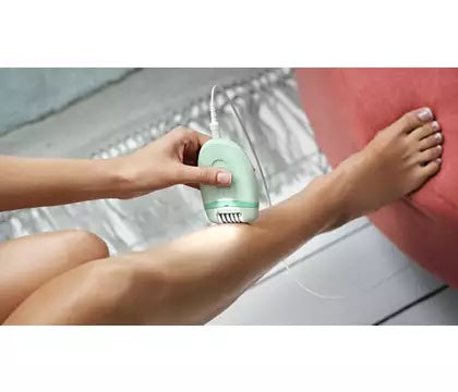 Billede af en person der bruger Philips BRE265/00 kompakt epilator med ledning på benet mens de sidder på en sofa i et lyst rum