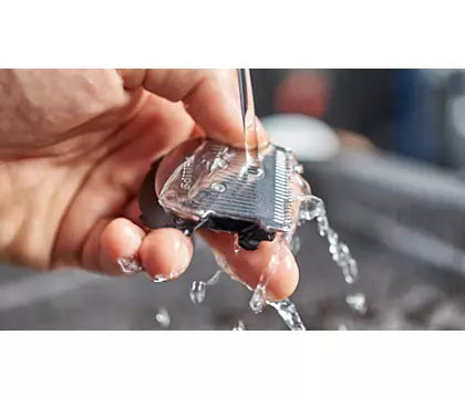 Hånd der holder en hårklipper mens vand sprøjter på klingen i en vask med fokus på rengøringsprocessen