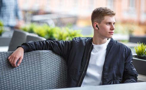 En ung mand sidder afslappet på en café med JVC HA-A8T-BU trådløse in-ear hovedtelefoner i sort farve, mens han kigger ud i det fjerne over en grå kurvestol og grønne planter.
