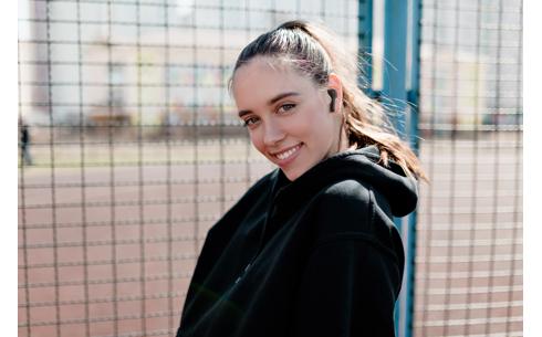 En ung kvinde i sort hoodie smiler mens hun står foran et gitterhegn med JVC HA-A8T-BU trådløse in-ear hovedtelefoner i ørerne