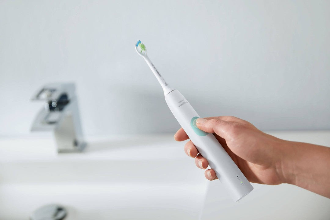 Elektrisk tandbørste Philips HX6807/24 Sonicare Protective Clean 4300 i en hånd med en grøn knap synlig og tandbørstehoveder i flere farver på toppen, med et moderne badeværelsesmiljø i baggrunden