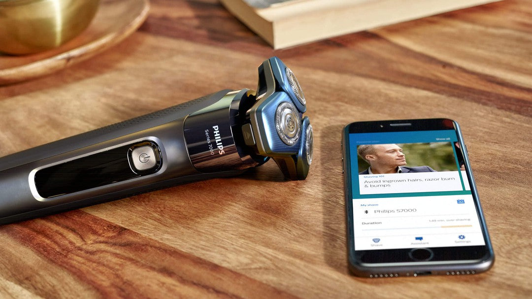 Elektrisk Philips S7782/50 Wet & Dryshaver ligger på et træbord ved siden af en smartphone der viser en app med information om barbering og pleje. Shaveren har et moderne design med tre roterende blade og en tænd/sluk knap, mens telefonen viser tips til at undgå indgroede hår og barberknopper.