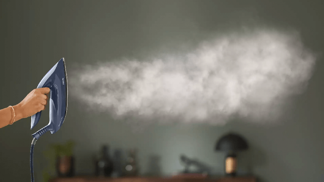 En person der holder en Philips Dampstation PSG7030/20 PerfectCare 7000 Series med damp der strømmer ud i en tåge i et stilrent rum med grøn væg og moderne indretning