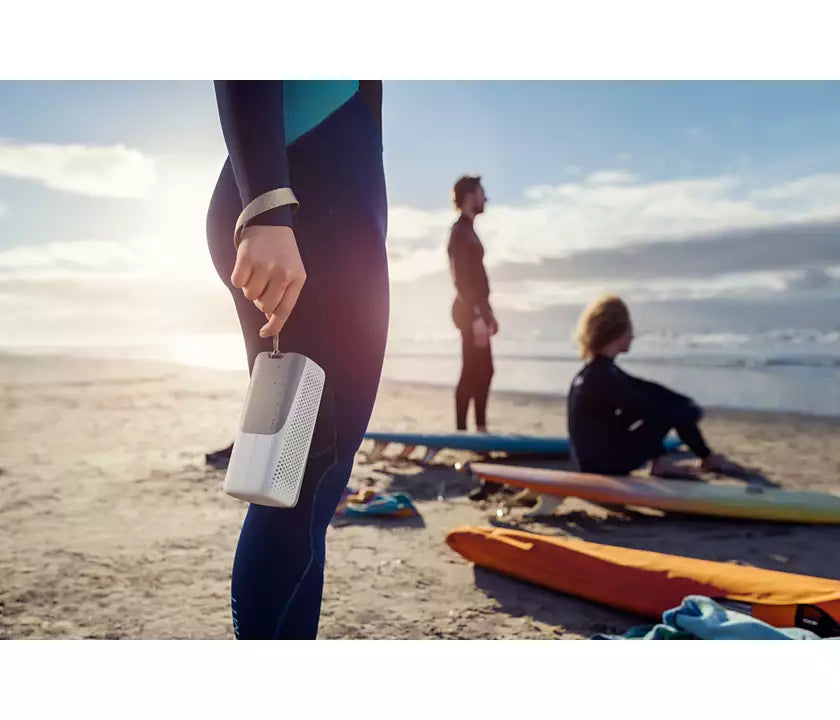 En person i våddragt holder en trådløs højttaler i hånden på en strand med surfbrætter i baggrunden og bølger, der slår mod kysten, i et solrigt vejr.