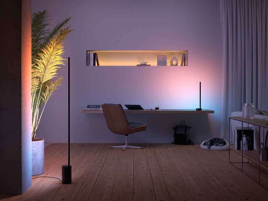 Billedet viser en moderne indendørs atmosfære med Philips HUE Signe gradient gulvlampe i sort placeret i et stilrent rum. Lampen kaster blødt, farvet lys på væggen og komplimenterer den minimalistiske indretning, der inkluderer en træbordplade med en ergonomisk stol og en plante. Der er også bøger og dekorative genstande i en hylde samt en hyggelig hundeseng i hjørnet.eteorisk og stilrent design.