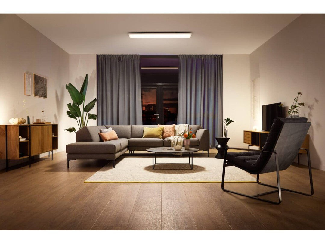 En moderne stue med Philips HUE Aurelle loftpanel monteret i loftet, blødt lys oplyser rummet, der er indrettet med en grå sofa, en sort lænestol og et rundt bord på en lys rug, planter dekorative elementer og kunstværker pryder væggene.