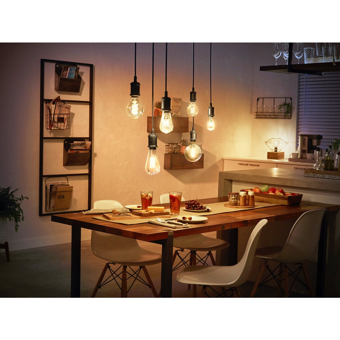 Billede af en moderne spisestue med en træbord dækket af snacks og drikkevarer omgivet af hvide stole flere filament pærer hængende fra loftet skaber en varm og indbydende atmosfære lampepærer har en Edison-stil design og giver blødt lys i rummet