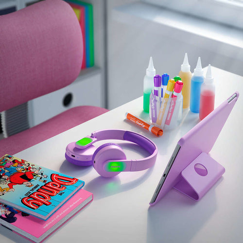 Trådløse on-ear hovedtelefoner til børn i lilla farve placeret på et bord sammen med en tablet, farverige tuscher, akrylmaling og en tegneserie.