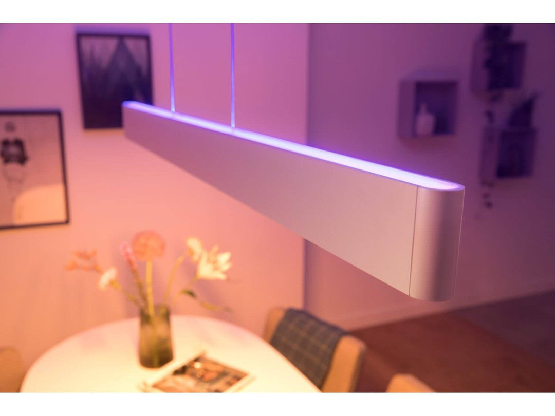 Philips HUE Ensis pendel i hvid farve hænger over et rundt bord med blomster i en vase ved siden af en bladansigt magasin belysning i bløde lilla nuancer i baggrunden
