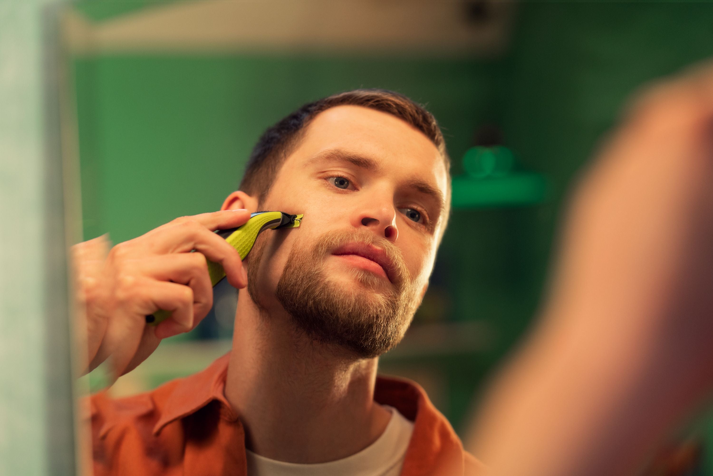 Mand med skæg barberer sit ansigt med Philips OneBlade 360 i et spejl omgivet af grønne vægge